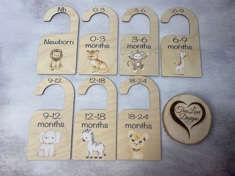 May include: Set of nine wooden hangers with animal illustrations and age ranges for baby clothes. The hangers are labeled with the following age ranges: Newborn, 0-3 months, 3-6 months, 6-9 months, 9-12 months, 12-18 months, 18-24 months. The animals include a lion, tiger, monkey, giraffe, elephant, zebra, and lion cub.