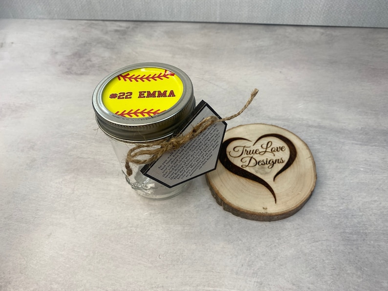 Puede incluir: Un tarro de cristal transparente con una tapa con tem&aacute;tica de softbol, con el texto "#22 EMMA". Una posavasos de madera con un dise&ntilde;o de coraz&oacute;n y las palabras "True Love Designs" est&aacute; al lado del tarro. Una etiqueta est&aacute; unida al tarro con hilo.