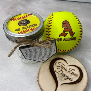 Puede incluir: Un juego de regalo con tem&aacute;tica de softbol que presenta una pelota de softbol amarilla con una "A" marr&oacute;n y "#9 ALLISON", un tarro con tapa amarilla y un posavasos de madera con "True Love Designs" y un dise&ntilde;o de coraz&oacute;n.
