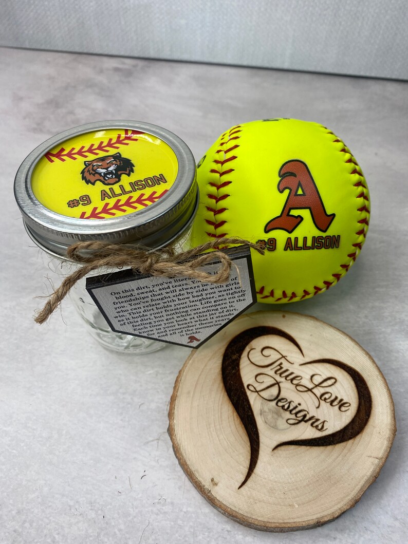 Puede incluir: Un juego de regalo con tem&aacute;tica de softbol que presenta una pelota de softbol amarilla con una "A" marr&oacute;n y "#9 ALLISON", un tarro con tapa amarilla y un posavasos de madera con "True Love Designs" y un dise&ntilde;o de coraz&oacute;n.