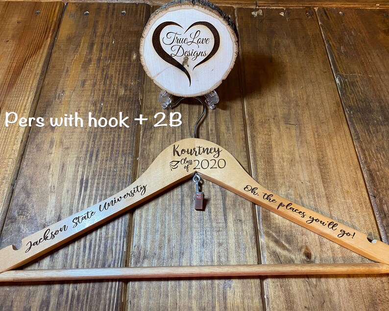 May include: A wooden clothes hanger with engraved text, including "Jackson State University" and "Kourtney Class of 2020." The hanger also features the phrase "Oh, the places you'll go!" and a small charm. A wooden disc with "True Love Designs" is above.