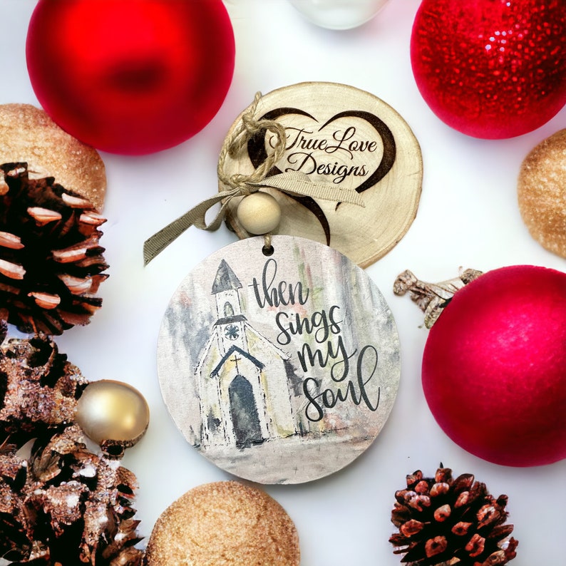May include: A round wooden ornament with a white church and the text "then sings my soul". The ornament is decorated with a brown twine bow and a small wooden heart with the text "True Love Designs".