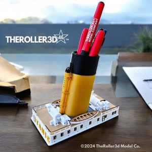 May include: A yellow and black 3D printed pen holder shaped like the iconic RMS Titanic. The pen holder has a red pen with the text "TheRoller3D Model Company" on it.