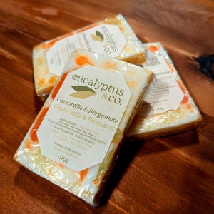 May include: Three bars of handmade soap with a white base and orange swirls. The soap is wrapped in clear plastic and labeled "eucalyptus & CO. Camomille & Bergamote Chamomile & Bergamot". The ingredients are listed on the label, including glycerin, shea butter, essential oil, mica powder, and more. The soap is made in Montreal and weighs 110 grams.