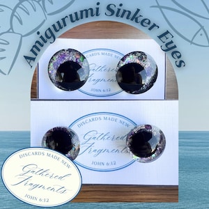 May include: Four circular amigurumi sinker eyes with black centers and glittery purple, silver, and green flecks. The eyes are mounted on two white display cards featuring the text Discards Made New Gathered Fragments John 6:12 in blue ink.