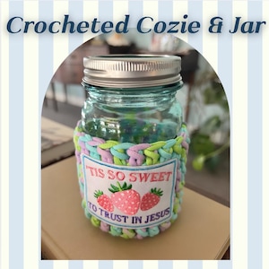 May include: A clear glass mason jar with a silver metal lid, encased in a pastel green, blue, and pink crocheted cozy. A rectangular white fabric patch is sewn onto the front, featuring three pink strawberries and the text 'TIS SO SWEET TO TRUST IN JESUS'.