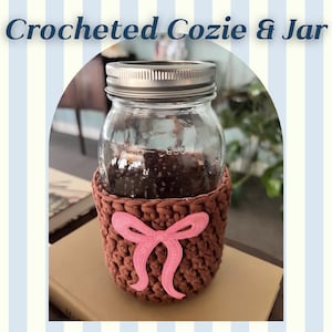 May include: A clear glass mason jar with a silver metal lid, encased in a brown crocheted cozy featuring a pink bow applique. The text Crocheted Cozie & Jar is displayed at the top of the image. The jar has a visible 800 milliliter marking.