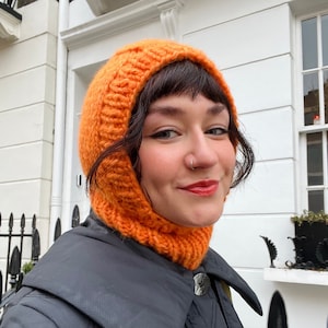 Può includere: Una persona indossa un cappello di lana arancione brillante con cappuccio e una giacca trapuntata nera. La persona è in piedi di fronte a un edificio bianco con una recinzione di ferro nero.