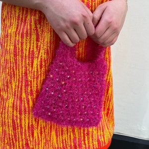 Può includere: Una borsetta rosa acceso e soffice con perline iridescenti sparse. La borsa è tenuta contro un vestito fantasia nei toni del giallo, arancione e rosso. L'abito presenta un motivo a righe verticali.