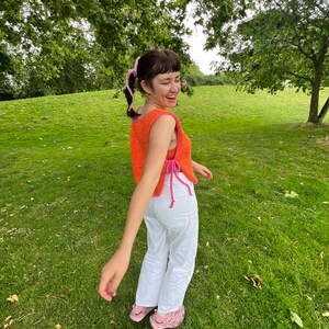 May include: A woman wearing a bright orange crochet top with pink accents and white trousers. She is standing in a grassy field with trees in the background.