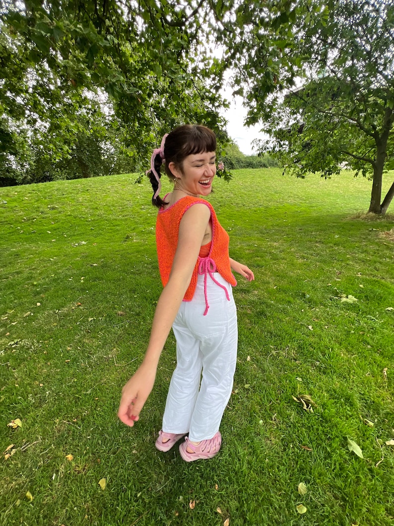 May include: A woman wearing a bright orange crochet top with pink accents and white trousers. She is standing in a grassy field with trees in the background.