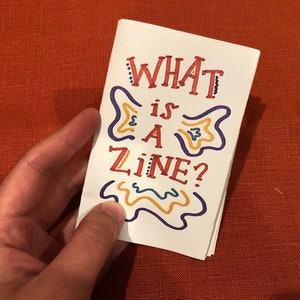 Puede incluir: Un pequeño folleto blanco con el texto "WHAT is A ZINE?" escrito en letras coloridas dibujadas a mano. Las letras están delineadas en morado y rellenas de amarillo y naranja.