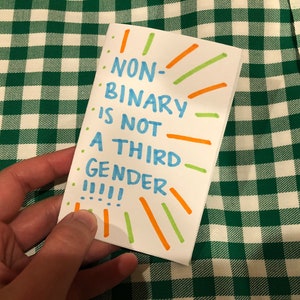 Puede incluir: Una tarjeta blanca con el texto "NO BINARIO NO ES UN TERCER GÉNERO !!!!" escrito en marcador azul. La tarjeta está decorada con líneas verdes y naranjas.