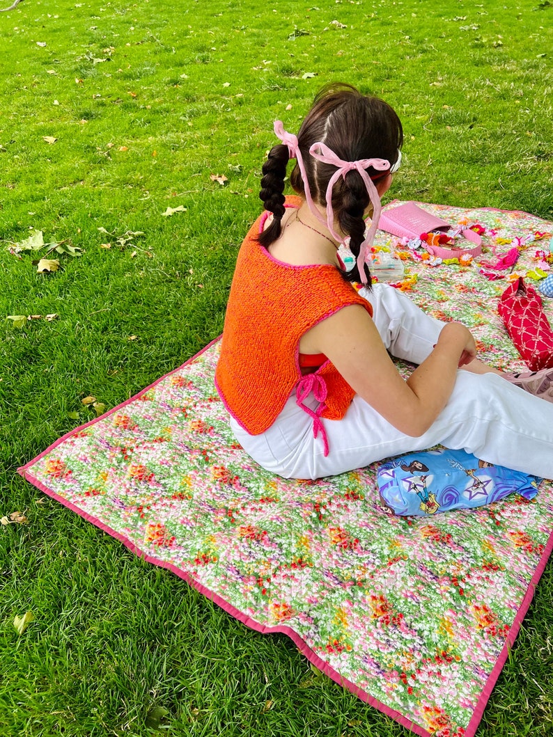 May include: A person is sitting on a floral patterned picnic blanket wearing a bright orange crocheted top and white trousers. Their hair is in pigtails with pink ribbons.