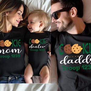 May include: Black t-shirts and a onesie with cookie-themed graphics. The shirts say "Cookie Mom" and "Cookie Daddy", and the onesie says "Cookie Dealer". All items include "Troop 1235".