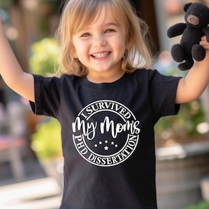 May include: A black t-shirt with a white circle graphic that says "I Survived My Mom's PhD Dissertation". A young child is wearing the shirt and smiling.