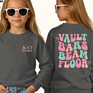 Puede incluir: Sudadera gris oscuro con dise&ntilde;o de gimnasia. La parte delantera presenta un peque&ntilde;o dise&ntilde;o bordado de gimnastas y la palabra "NAME". La parte trasera muestra las palabras "VAULT BARS BEAM FLOOR" en una fuente retro, con corazones.