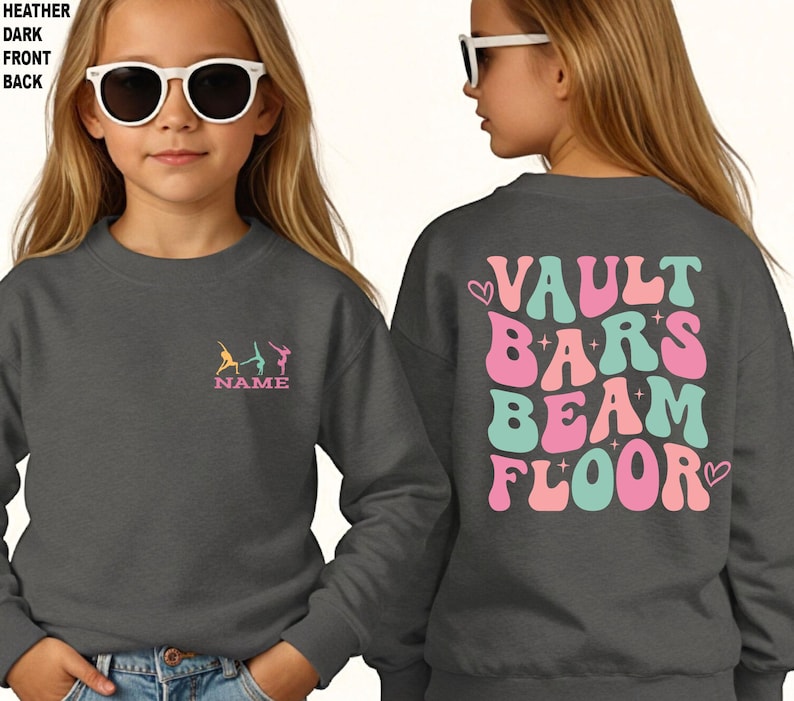 Puede incluir: Sudadera gris oscuro con dise&ntilde;o de gimnasia. La parte delantera presenta un peque&ntilde;o dise&ntilde;o bordado de gimnastas y la palabra "NAME". La parte trasera muestra las palabras "VAULT BARS BEAM FLOOR" en una fuente retro, con corazones.