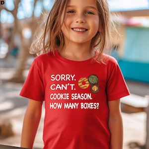May include: A red t-shirt with the text "SORRY. CAN'T. COOKIE SEASON. HOW MANY BOXES?" and cookie graphics. The shirt is short-sleeved and worn by a young person.