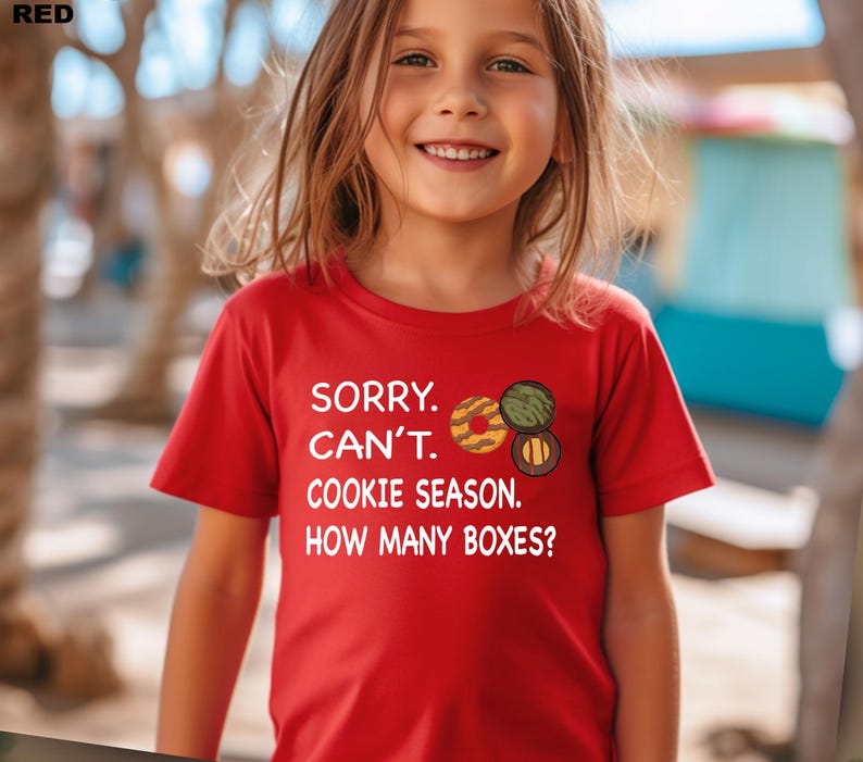 May include: A red t-shirt with the text "SORRY. CAN'T. COOKIE SEASON. HOW MANY BOXES?" and cookie graphics. The shirt is short-sleeved and worn by a young person.