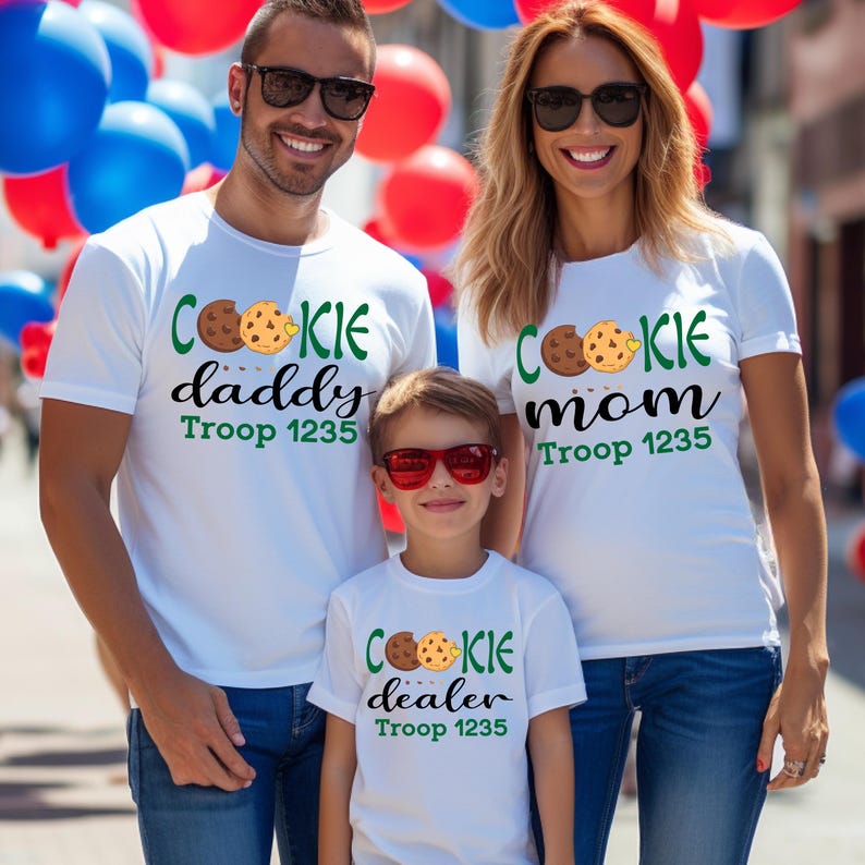 May include: Three people wearing white t-shirts with cookie-themed designs. The shirts say "Cookie Daddy," "Cookie Mom," and "Cookie Dealer," with cookie graphics. The text "Troop 1235" is also printed on the shirts.