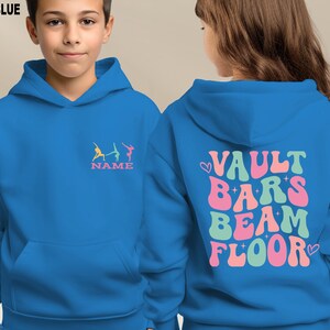 Puede incluir: Sudadera con capucha azul real con bolsillo delantero y capucha. La parte delantera presenta un dise&ntilde;o de gimnasia colorido con la palabra "NAME" debajo. La parte trasera muestra las palabras "VAULT BARS BEAM FLOOR" en una fuente retro.