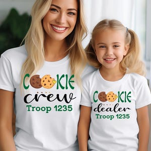 May include: White t-shirts with matching designs. One shirt says "COOKIE crew" and the other says "COOKIE dealer". Both shirts have a cookie graphic and "Troop 1235" in green. The shirts are for a mother and daughter.