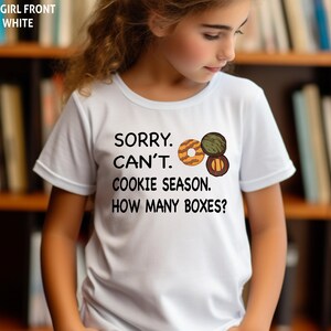 May include: White t-shirt with the text "SORRY. CAN'T. COOKIE SEASON. HOW MANY BOXES?" and a graphic of three cookies. The shirt is short-sleeved.