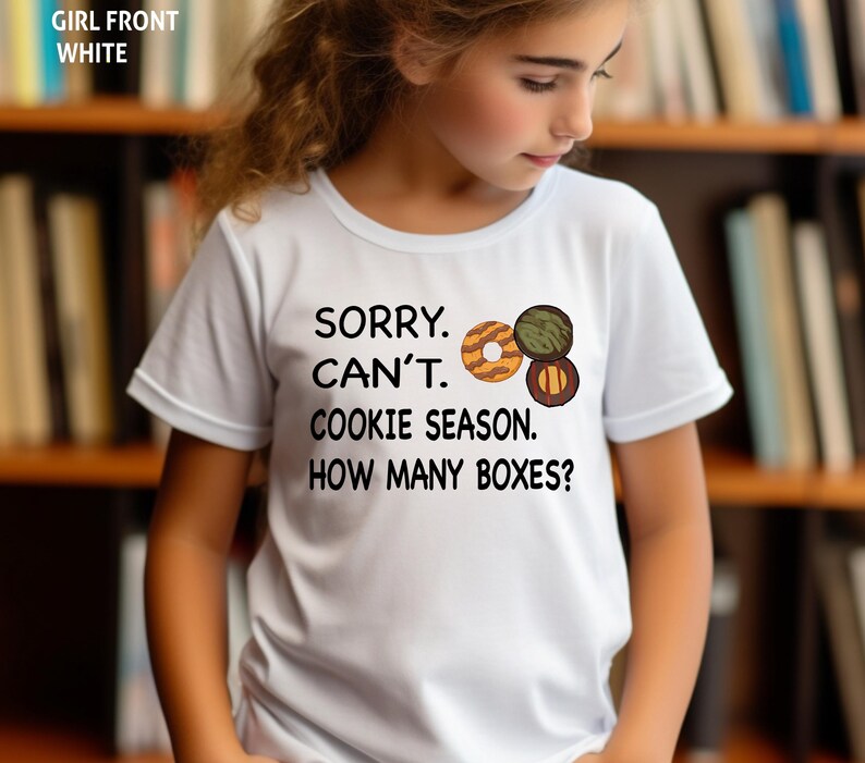 May include: White t-shirt with the text "SORRY. CAN'T. COOKIE SEASON. HOW MANY BOXES?" and a graphic of three cookies. The shirt is short-sleeved.