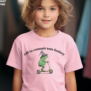 May include: Light pink t-shirt with the text "Off to commit tom foolery." The shirt features a green frog wearing a wizard hat and riding a scooter. The frog is the central graphic on the shirt.