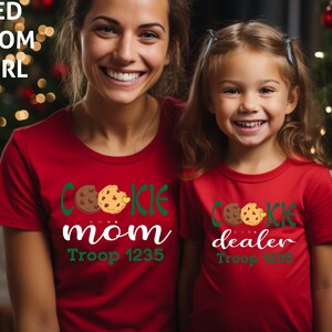 May include: Two people wearing matching red t-shirts with cookie-themed designs. One shirt says "Cookie Mom" and the other says "Cookie Dealer". Both shirts have a cookie graphic and the text "Troop 1235". The text "RED MOM GIRL" is in the top left corner.