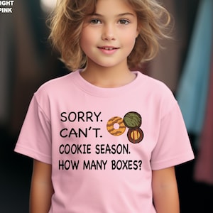 May include: Light pink t-shirt with the text "SORRY. CAN'T. COOKIE SEASON. HOW MANY BOXES?" and a graphic of three cookies. The shirt is worn by a young person.