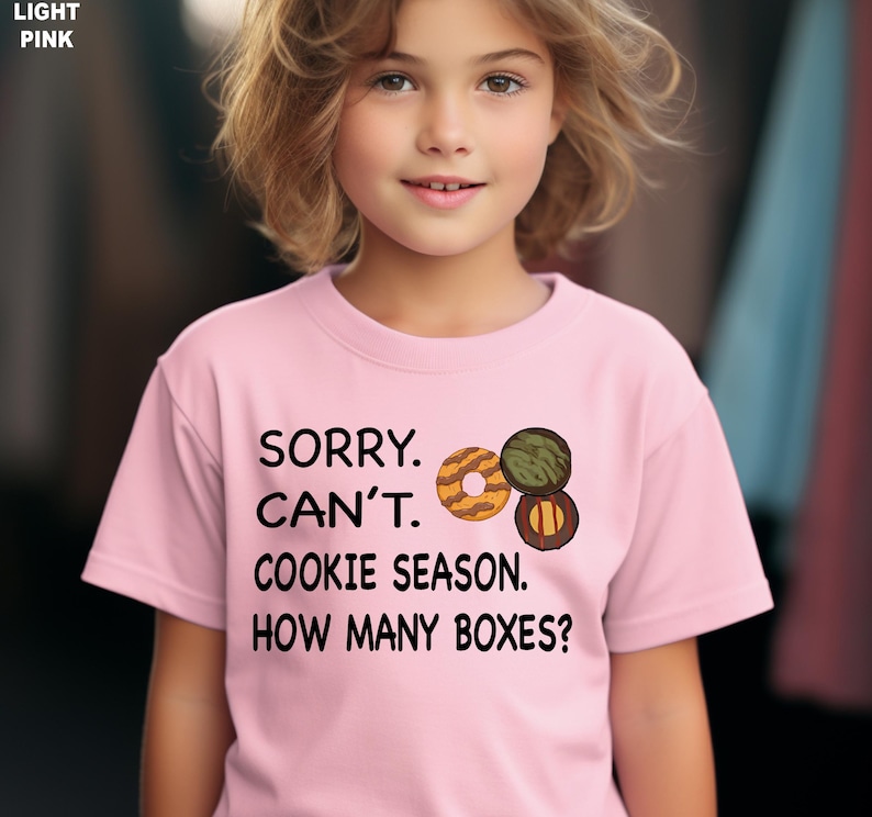 May include: Light pink t-shirt with the text "SORRY. CAN'T. COOKIE SEASON. HOW MANY BOXES?" and a graphic of three cookies. The shirt is worn by a young person.