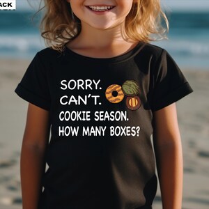May include: Black t-shirt with the text "SORRY. CAN'T. COOKIE SEASON. HOW MANY BOXES?" and illustrations of cookies. The shirt is worn by a child on a beach.