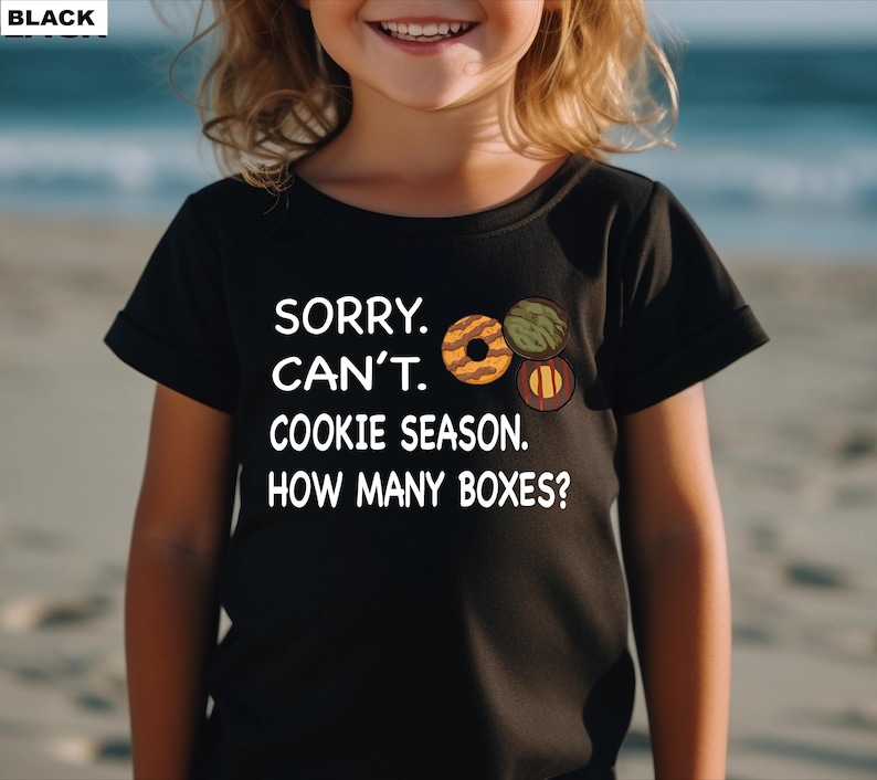 May include: Black t-shirt with the text "SORRY. CAN'T. COOKIE SEASON. HOW MANY BOXES?" and illustrations of cookies. The shirt is worn by a child on a beach.