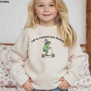May include: Beige sweatshirt with the text "Off to commit tom foolery." The graphic features a green frog wearing a wizard hat and riding a scooter. The sweatshirt is a neutral color.