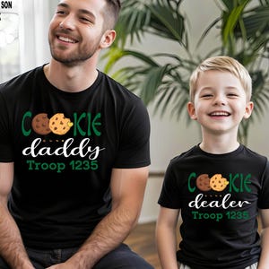 May include: Black t-shirts with cookie-themed graphics. One shirt says "Cookie Daddy" and the other says "Cookie Dealer", both with "Troop 1235". The shirts feature a chocolate chip cookie design in green, brown, and yellow.