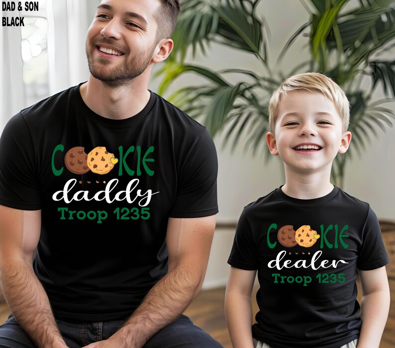 May include: Black t-shirts with cookie-themed graphics. One shirt says "Cookie Daddy" and the other says "Cookie Dealer", both with "Troop 1235". The shirts feature a chocolate chip cookie design in green, brown, and yellow.