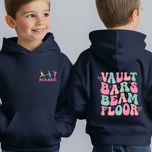 Puede incluir: Sudadera con capucha azul marino. La parte delantera muestra figuras de gimnastas en amarillo, turquesa y rosa, con la palabra "NAME" debajo. La parte trasera tiene las palabras "VAULT BARS BEAM FLOOR" en rosa y turquesa, con corazones.