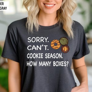 May include: Heather dark gray t-shirt with the text "SORRY. CAN'T. COOKIE SEASON. HOW MANY BOXES?" and illustrations of three cookies. The shirt is a dark gray color.