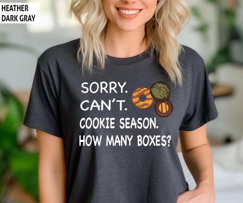 May include: Heather dark gray t-shirt with the text "SORRY. CAN'T. COOKIE SEASON. HOW MANY BOXES?" and illustrations of three cookies. The shirt is a dark gray color.