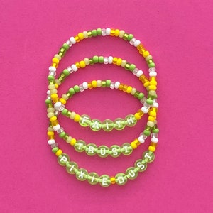 May include: Four beaded bracelets with the words "Faith", "Trust", and "Pixie Dust" spelled out in white beads on a green background. The bracelets are layered on top of each other and are made with green, yellow, and white beads.
