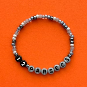Könnte beinhalten: Ein graues und weißes Perlenarmband mit den Worten "12 Parsecs" in schwarzen Buchstaben.