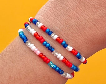 May include: Three beaded bracelets in red, white, and blue colors. The bracelets are stacked on a person's wrist.