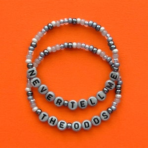 Könnte beinhalten: Zwei Perlenarmbänder mit silbernen und weißen Perlen. Die Armbänder buchstabieren "Never Tell The Odds" und "Me".