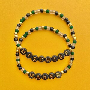 May include: Two beaded bracelets with black and gold beads. The top bracelet spells out "MISCHIEF" and the bottom bracelet spells out "MAKER".
