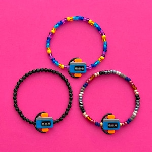 May include: Three beaded bracelets with colorful beads and a charm in the shape of a small robot. The robot charm is blue, gray, and orange.