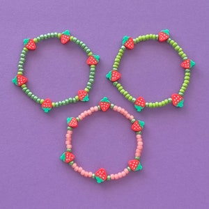 Puede incluir: Tres pulseras con dijes de fresa. Las pulseras están hechas con pequeñas cuentas verdes, rosadas y blancas.