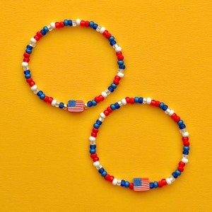 Puede incluir: Dos pulseras de cuentas rojas, blancas y azules con un pequeño dije de bandera estadounidense en cada una.