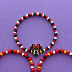 May include: Three beaded bracelets with a Minnie Mouse theme. The bracelets feature red, black, white, and yellow beads. One bracelet has a Minnie Mouse charm. The bracelets are on a purple background.